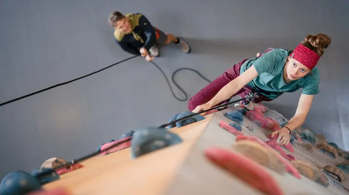 Eine Frau klettert in einer Kletterhalle, ihr Partner sichert sie. | © DAV / Julian Rohn